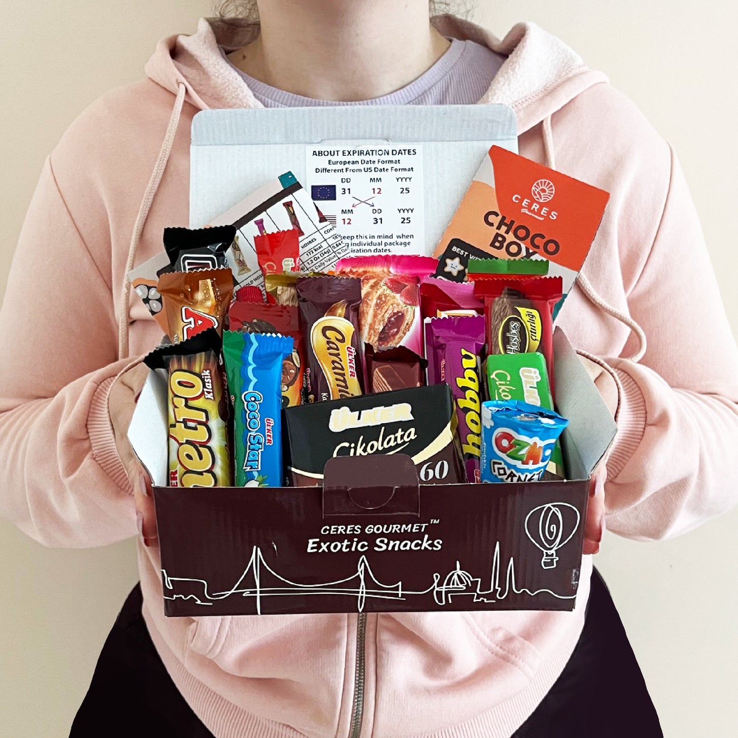 A person in a pink hoodie holds a Ceres Gourmet International Chocolate Variety Box (21 pieces) and a pamphlet with expiration date information.