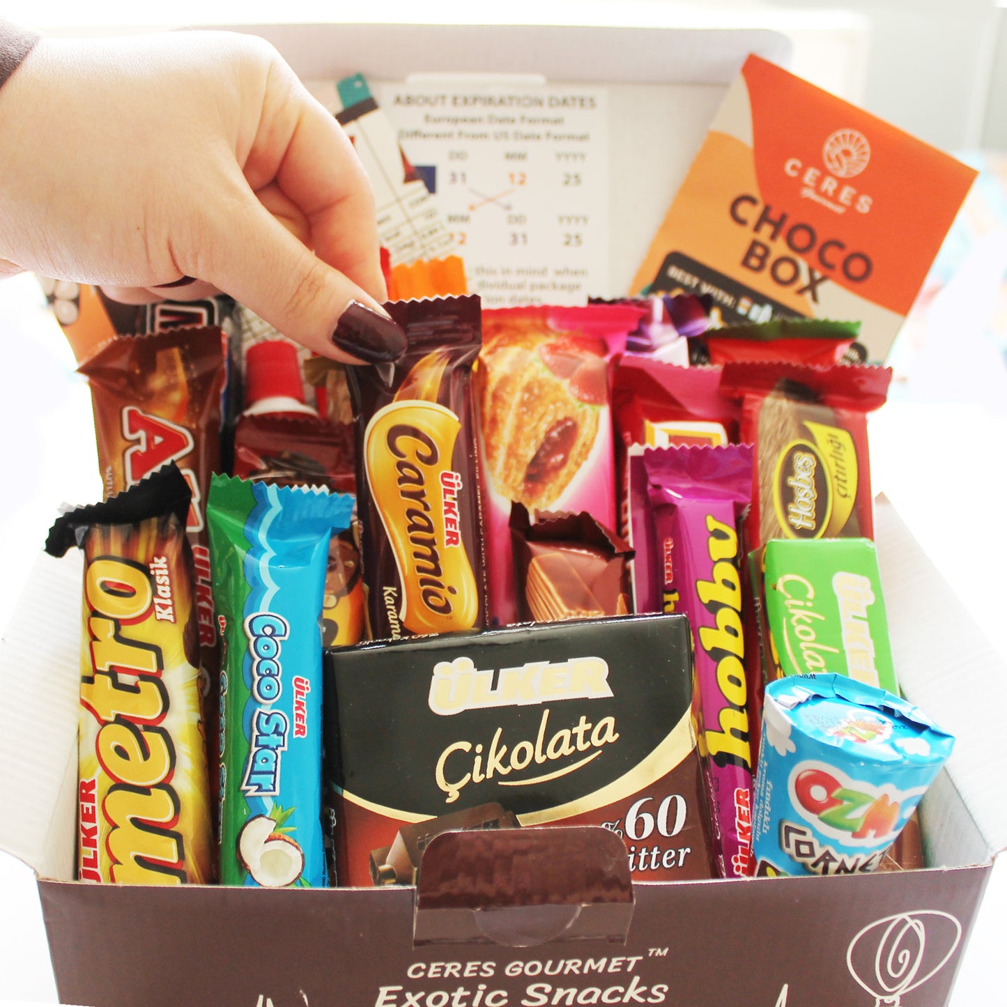 A hand with painted nails reaches into a Ceres Gourmet International Chocolate Variety Box 🍫21 Pieces, filled with assorted treats and Turkish candies like Metro, Caramio, Coco Star, and Ülker. A snack guide is also visible in the background.