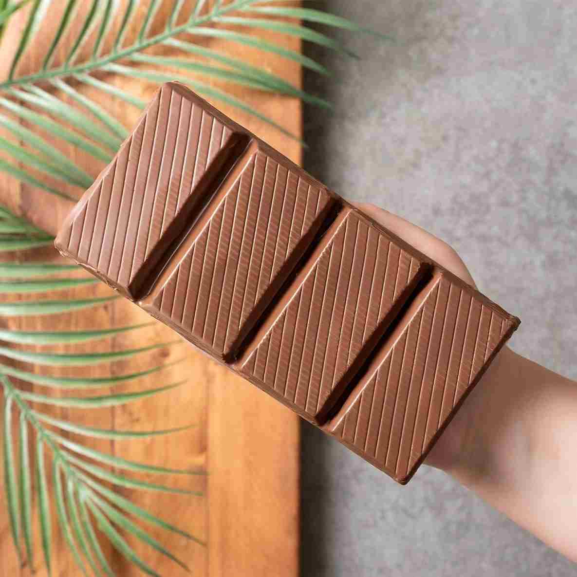 A hand holds the Ceres Gourmet Viral Halloween Dubai Chocolate Bar, 75% Pistachio (7 oz)—a large, handcrafted treat with four segments and pistachio cream, set on wood with a palm leaf and gray backdrop.