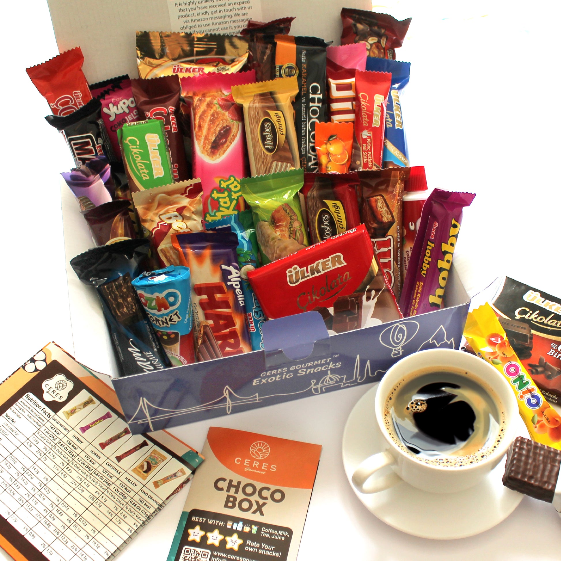 An open International Chocolate Variety Box 31 Pieces đ« by Ceres Gourmet sits next to a cup of coffee, a tasting guide with scoring chart, and a CHOCObox info card, all arranged on a white surface.