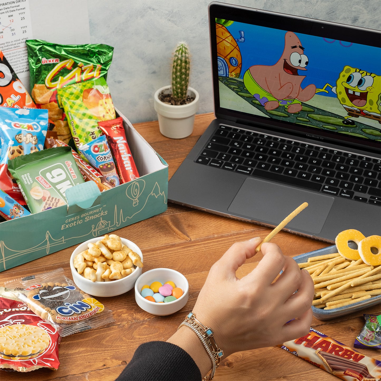 A hand holds a snack stick at a desk with Ceres Gourmets Snack Boxes from Around the World—22 Delicious Snacks—plus global treats, a laptop streaming SpongeBob SquarePants, a potted cactus, and papers in the background.