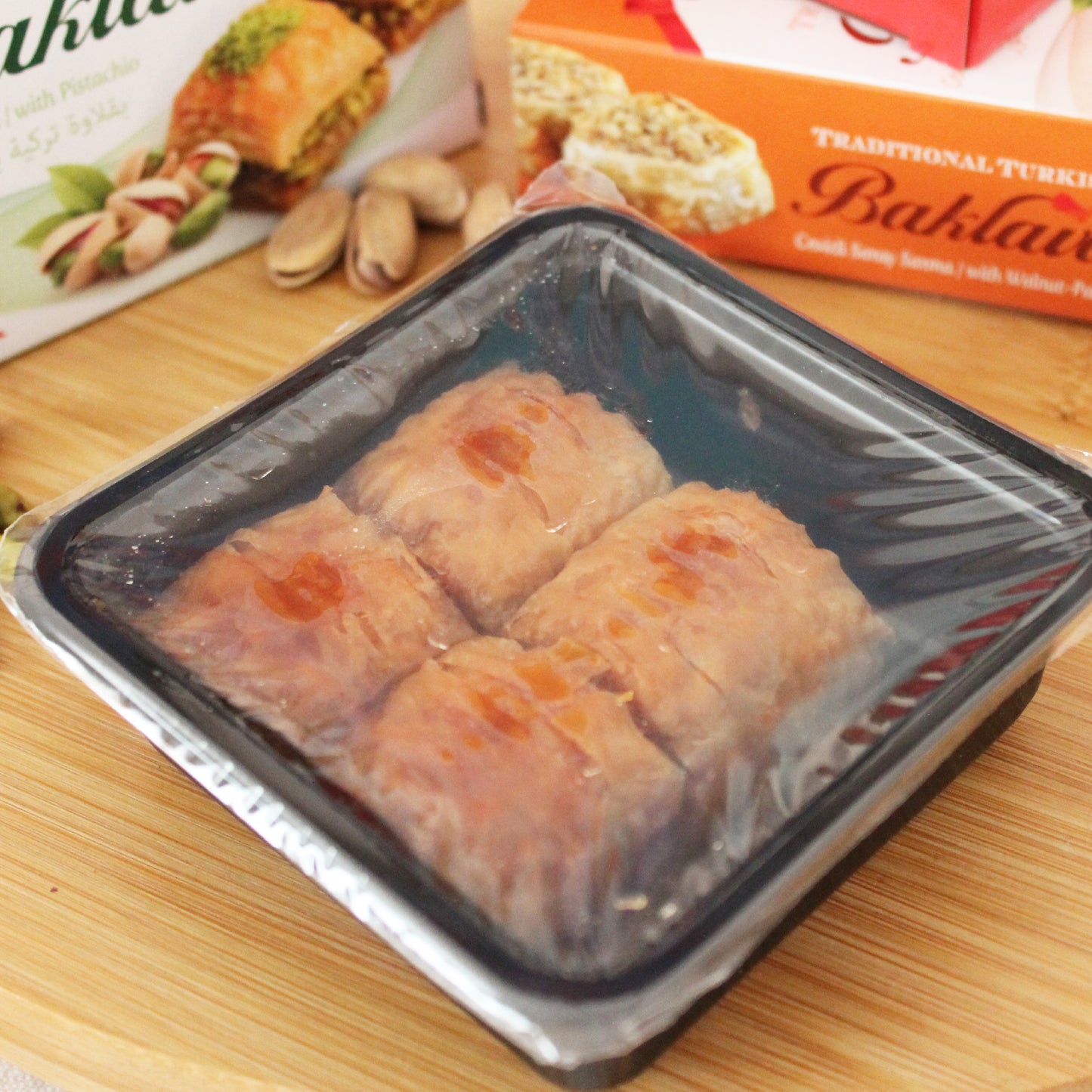 A Ceres Gourmet Assorted Turkish Baklava Variety Gift Set, Pistachio and Walnut 4 Pack, is displayed in a plastic container on a wooden surface, with baklava gift boxes and pistachios in the background.