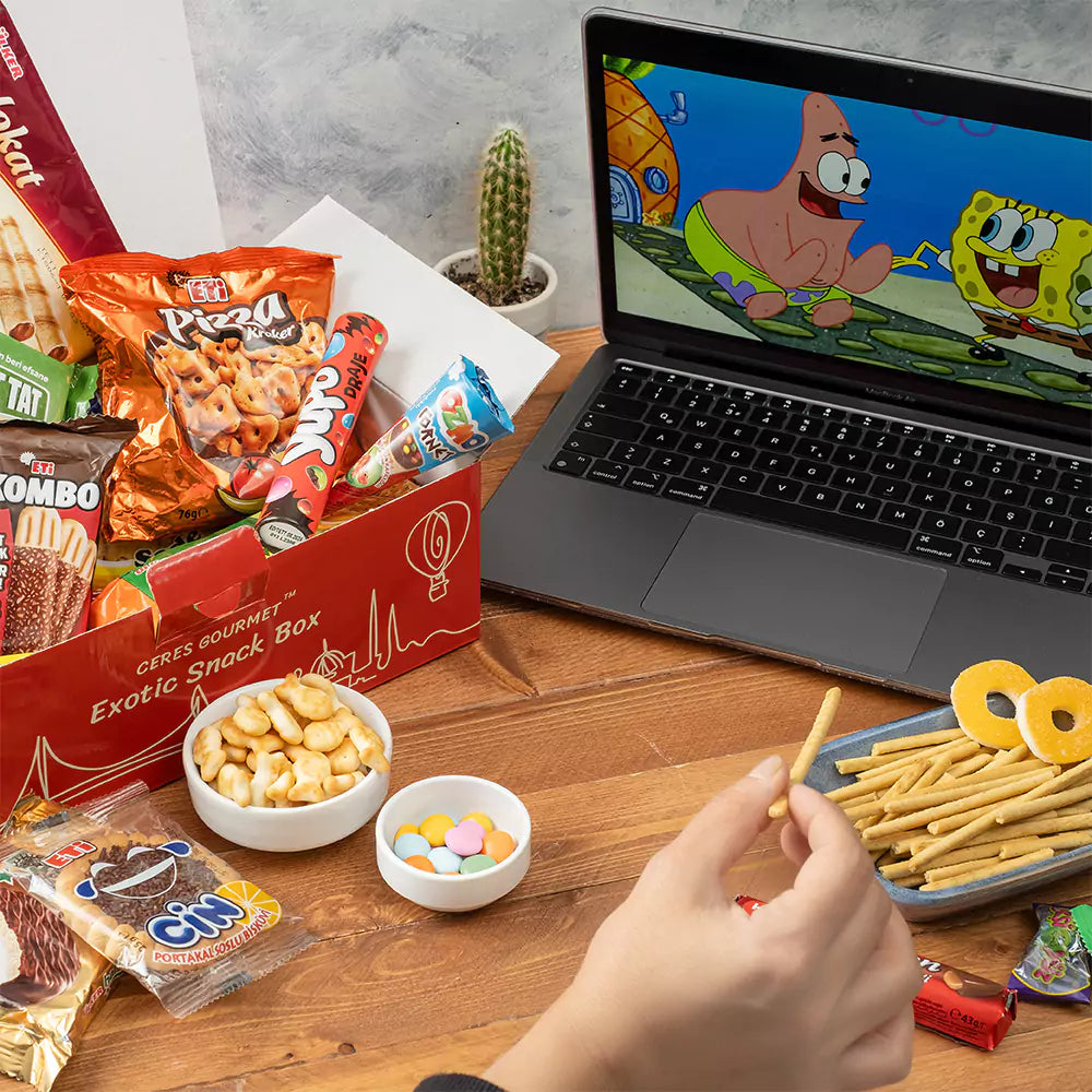A hand grabs a treat from the Ceres Gourmet 50-Pack International Snack Box - Assorted Treats on a wooden table with snacks, a red box, and an open laptop showing SpongeBob, while a small cactus decorates the background.