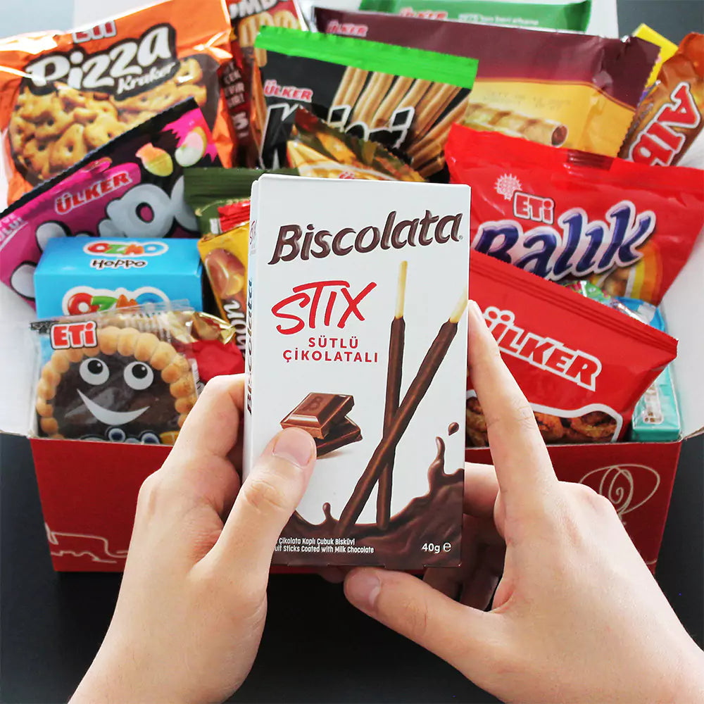 A person holds a box of Biscolata Stix in front of a Ceres Gourmet 50-Pack International Snack Box, filled with assorted colorful Turkish treats—an ideal option for employee appreciation or corporate gifting.