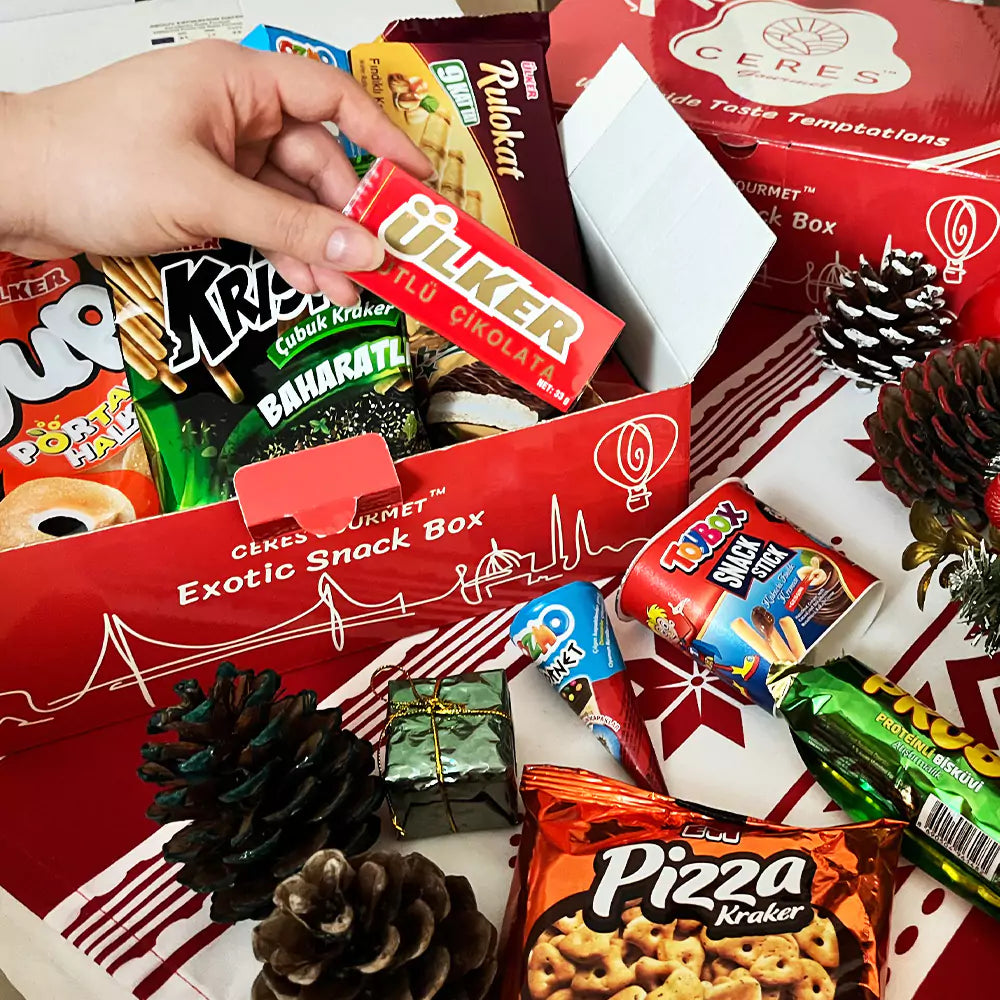 A hand reaches into a Ceres Gourmet 50-Pack International Snack Box – Assorted Treats, ideal for corporate gifting or employee appreciation. Pinecones and snacks decorate the festive table around the bright red box.