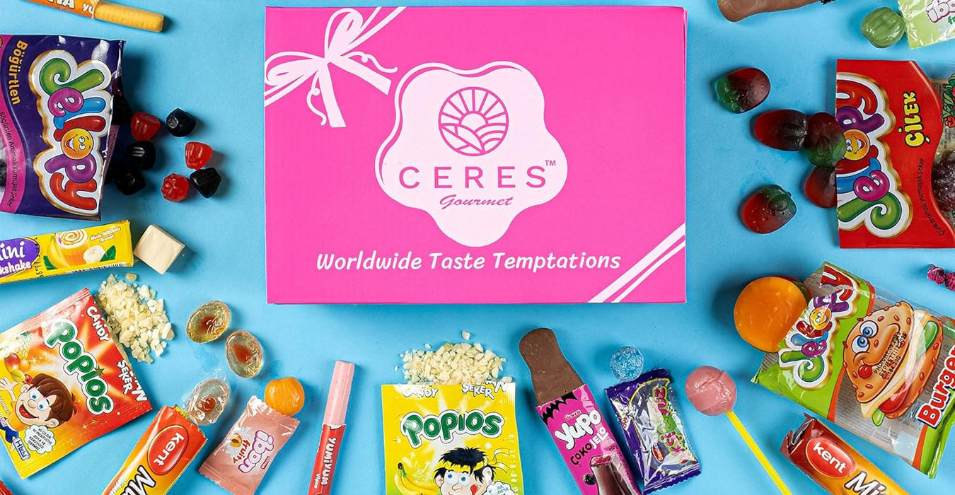 A pink Ceres Gourmet box sits in the center, surrounded by various colorful international candies and snacks on a blue background. The box reads “Worldwide Taste Temptations” and is decorated with a white ribbon graphic.