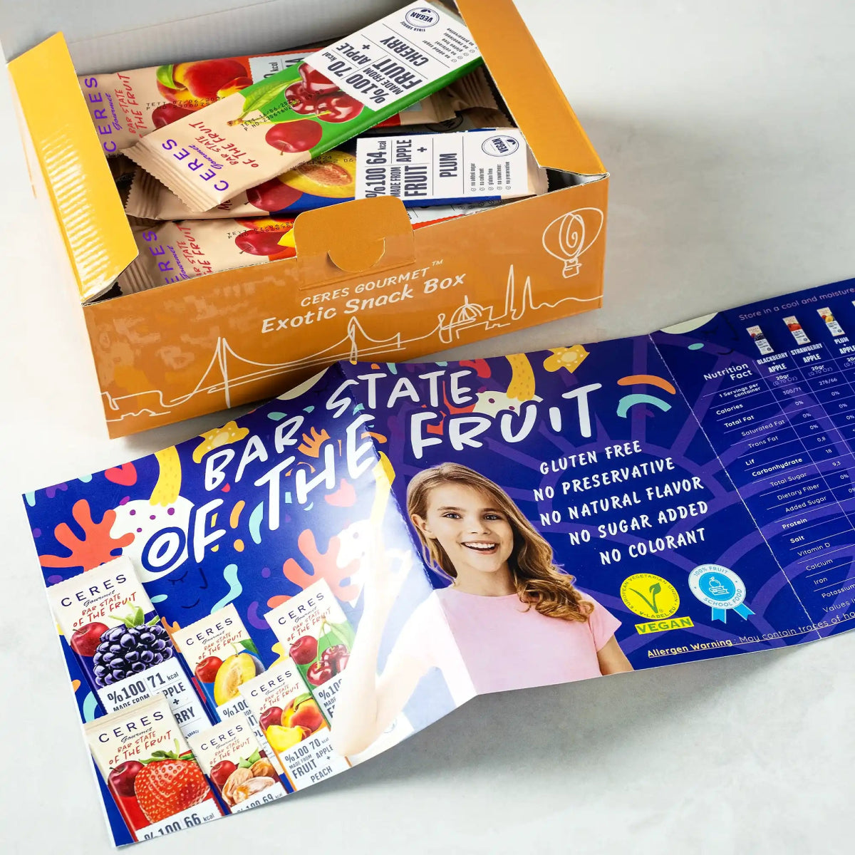 A partially open Fruit Strips Variety Pack by Ceres Gourmet sits next to a colorful brochure with fruit images, nutrition info, and a smiling woman in pink, highlighting gluten-free bars, vegan claims, and no added sugar.