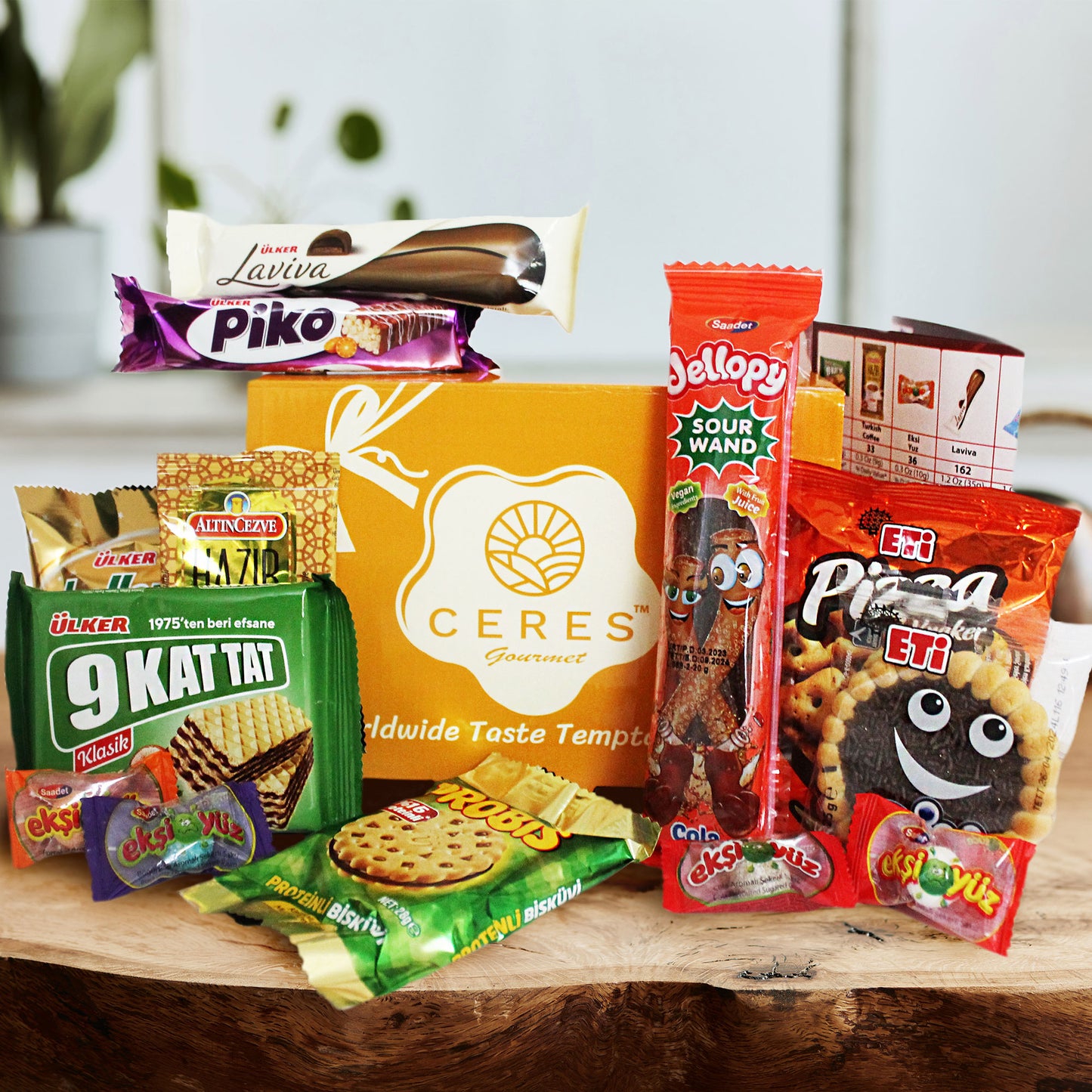 A vibrant array of packaged cookies, wafers, crackers, and candy surrounds the yellow Ceres Gourmet International Exotic Treats Box—featuring 15 premium foreign rare snacks—on a wooden surface with blurred plants in the background.