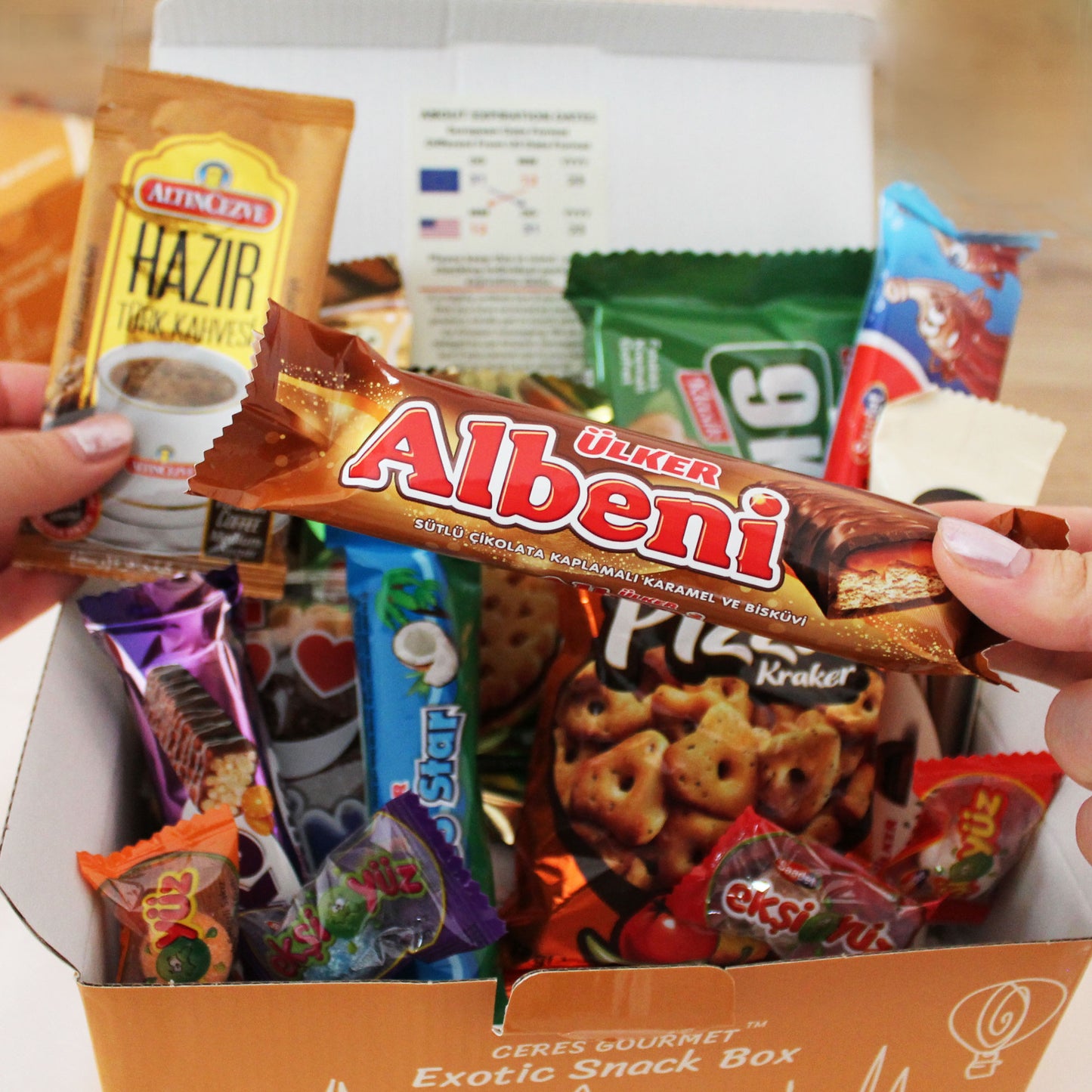 A person holds a Ülker Albeni bar above an open Ceres Gourmet International Exotic Treats Box, featuring 15 premium foreign rare snacks like coffee, pretzels, and assorted colorful delights.