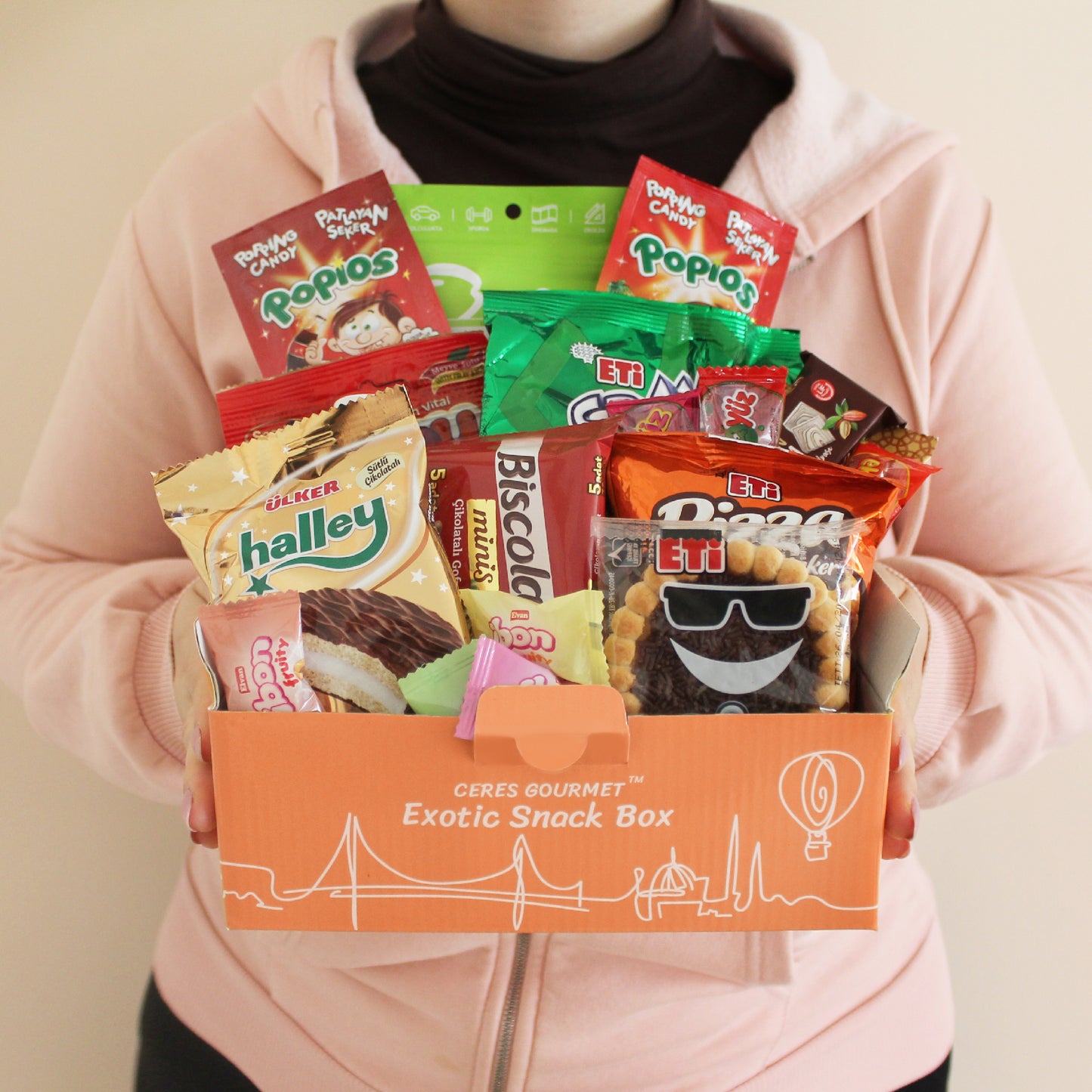 A person in a pink hoodie holds an orange box labeled Ceres Gourmet International Snack Box with 12 different snacks and 15+ pieces, filled with assorted colorful treats and rare foreign candies for a global snacking experience.
