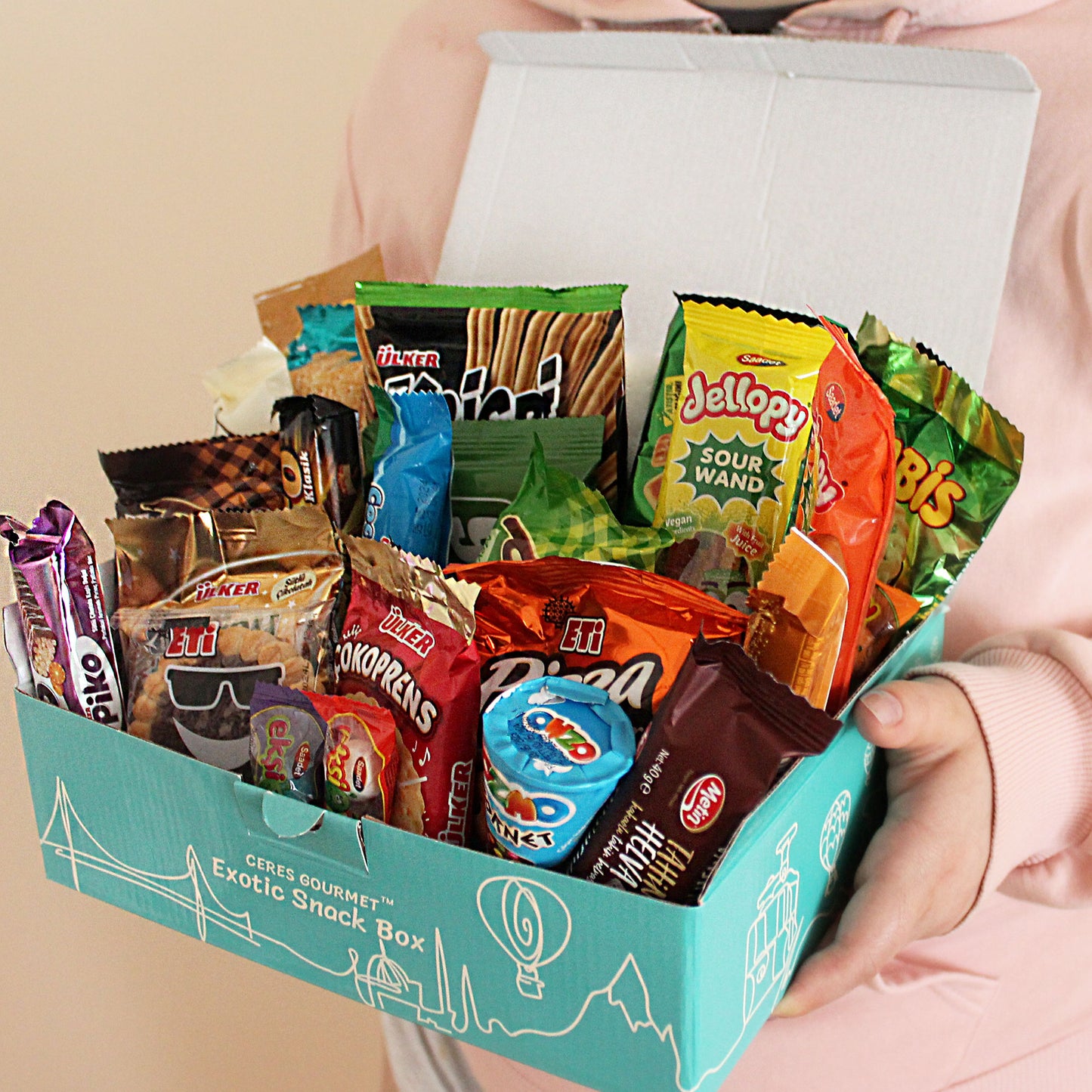 A person holds an open turquoise box labeled Exotic Snack Box, filled with a variety of colorful packaged snacks, including cookies, wafers, chips, and candy.