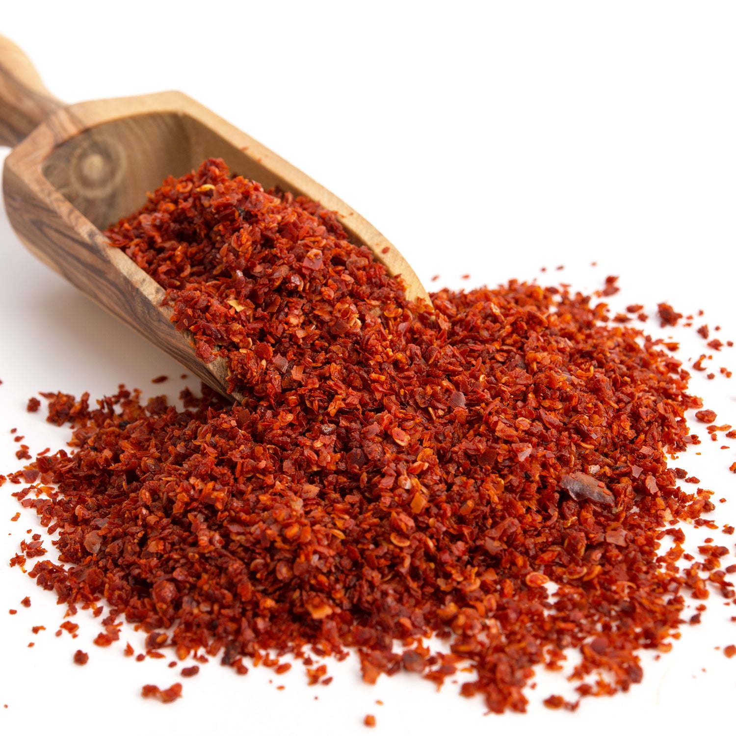A wooden scoop pours Ceres Gourmet Premium Aleppo Pepper 7 Oz, freshly ground for exquisite flavor and mild heat, onto a white surface.