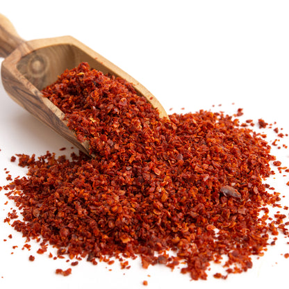 A wooden scoop pours Ceres Gourmet Premium Aleppo Pepper 7 Oz, freshly ground for exquisite flavor and mild heat, onto a white surface.