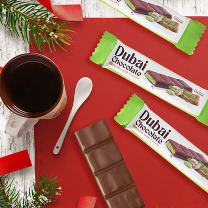 A red table displays Ceres Gourmet Dubai Chocolate Pistachio Kunafa Bars (one unwrapped), a coffee cup with a spoon, and pine branches with festive red ribbons on a white wooden background.