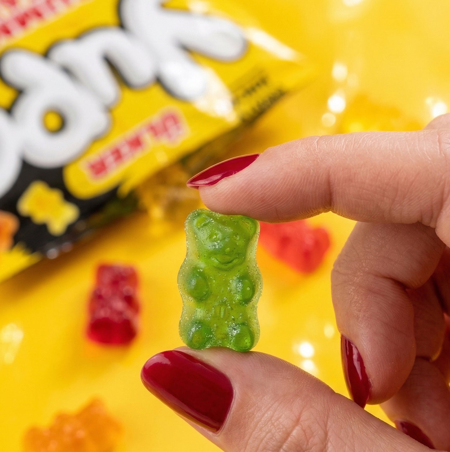 Green gummy bear held by a hand with red nail polish, against a yellow background with candy packaging.