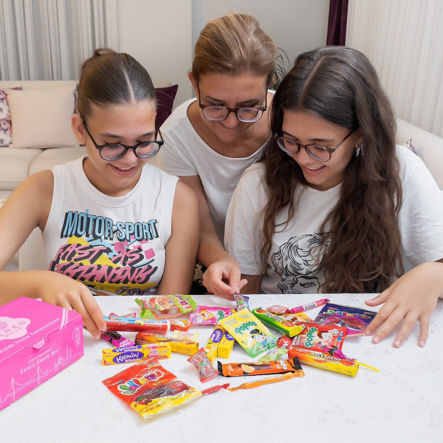 Three people with long hair and glasses smile as they pick from the Ceres Gourmet Candy Snacks Box—40 rare, unique international candies—enjoying sweet surprises and discovering new flavors in a bright, cozy room.