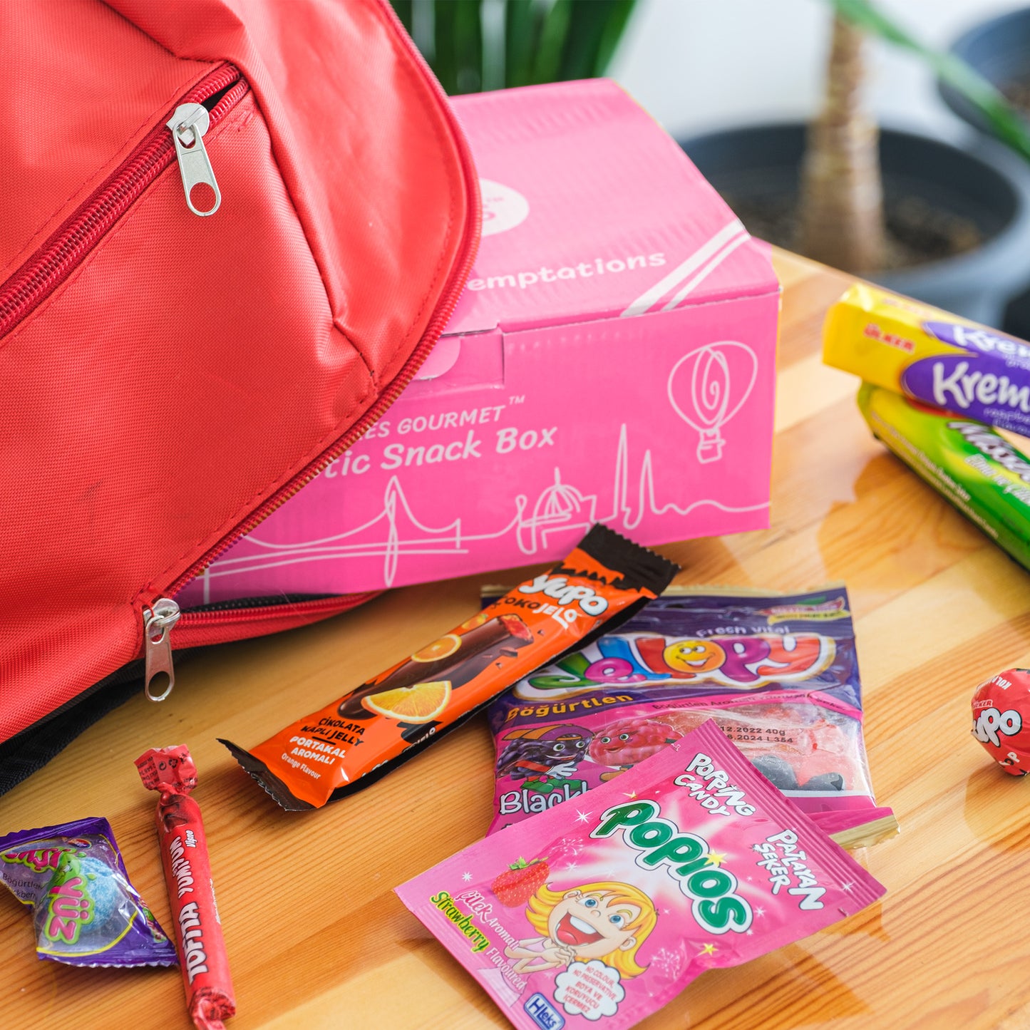 A red backpack and a pink snack box sit on a wooden table, surrounded by Ceres Gourmet’s Candy Snacks Box Foreign Rare Unique 40 Mystery Candy, featuring lollipops, chewy bars, and playful packets.