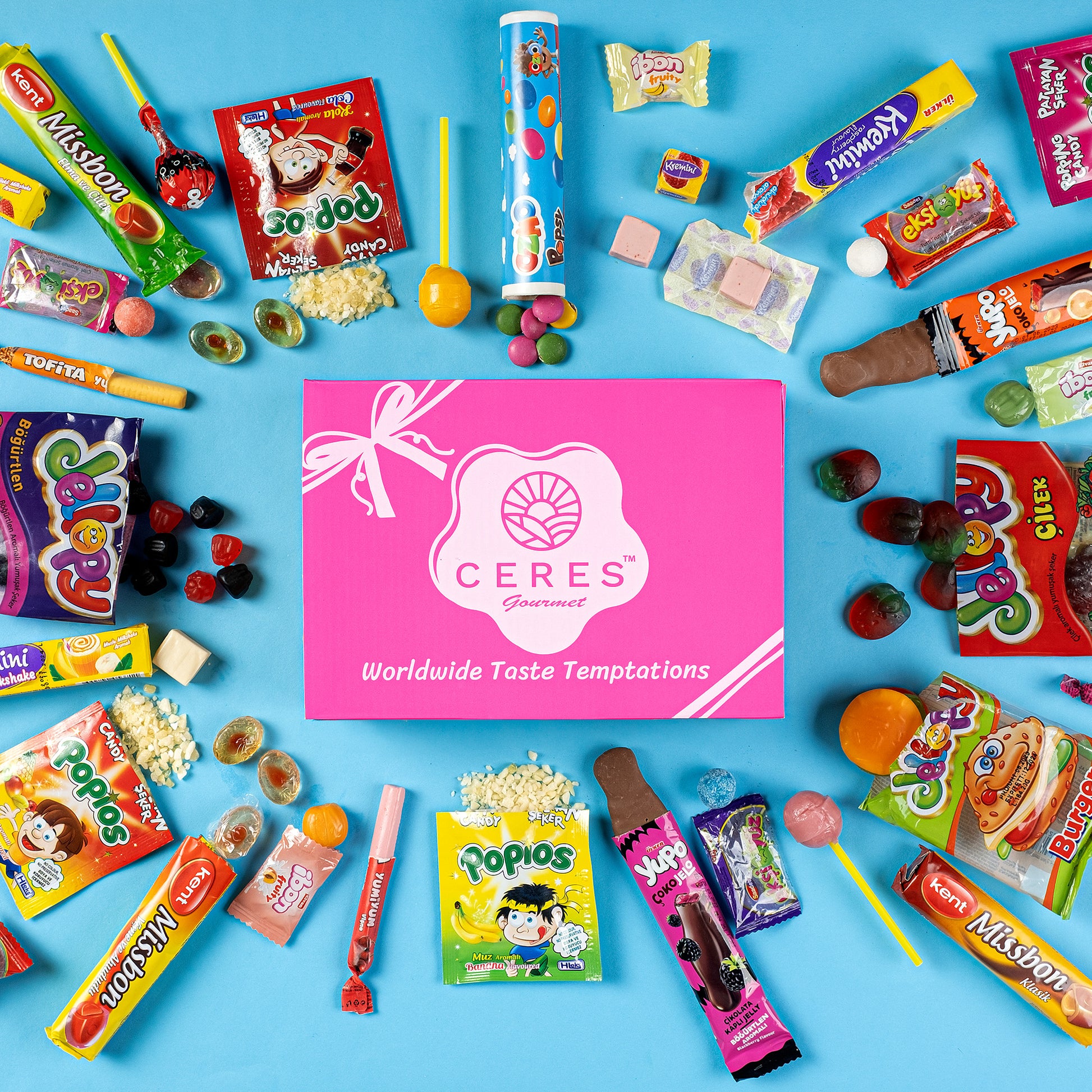 A flat lay of assorted colorful international candies and snacks surrounds a pink Ceres Gourmet Candy Snacks Box Foreign Rare Unique 40 Mystery Candy on a blue background.