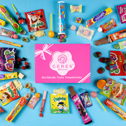 A flat lay of assorted colorful international candies and snacks surrounds a pink Ceres Gourmet Candy Snacks Box Foreign Rare Unique 40 Mystery Candy on a blue background.
