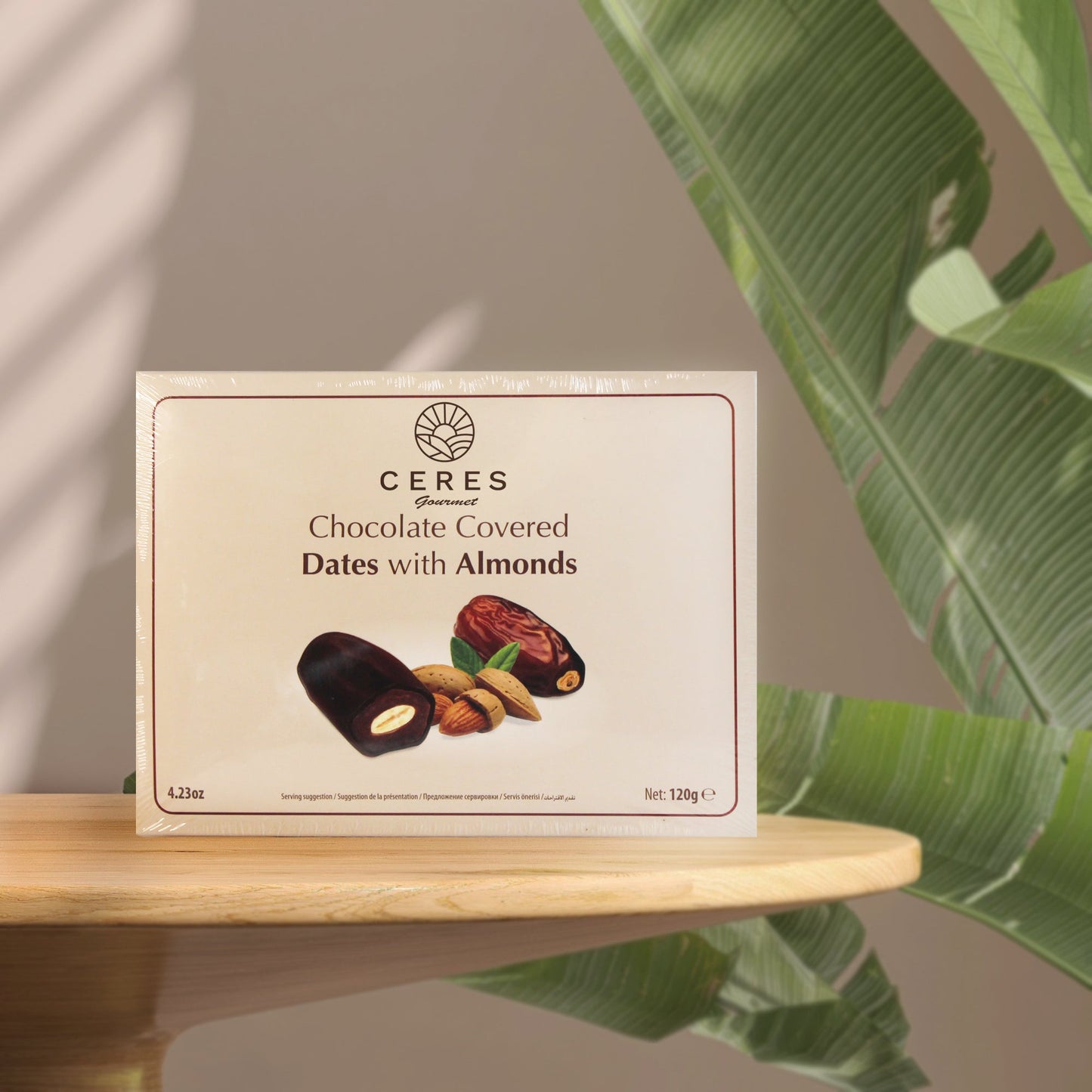 A box of Ceres Gourmet Dark Chocolate Coated Medjool Dates Stuffed with Almonds (2 packs, 8.46 oz) sits on a round wooden table, surrounded by green leaves, featuring images of these delicious treats and their almond-filled centers.