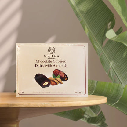 A box of Ceres Gourmet Dark Chocolate Coated Medjool Dates Stuffed with Almonds (2 packs, 8.46 oz) sits on a round wooden table, surrounded by green leaves, featuring images of these delicious treats and their almond-filled centers.
