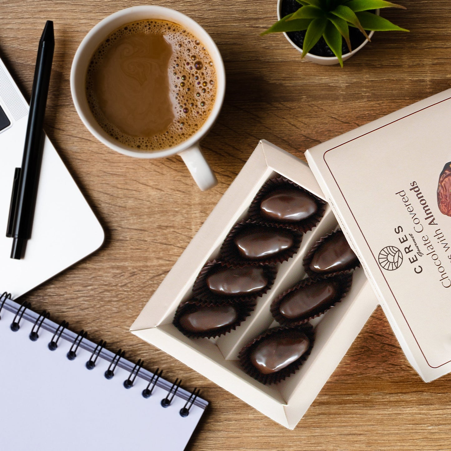 A pack of Ceres Gourmet Dark Chocolate Coated Medjool Dates Stuffed with Almonds (2 packs, 8.46 oz) sits by coffee, a notebook, pen, laptop, and a plant—a perfect healthy workspace snack.