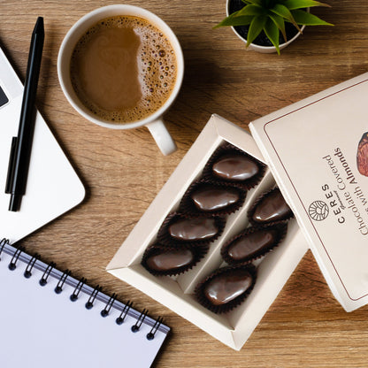 A pack of Ceres Gourmet Dark Chocolate Coated Medjool Dates Stuffed with Almonds (2 packs, 8.46 oz) sits by coffee, a notebook, pen, laptop, and a plant—a perfect healthy workspace snack.