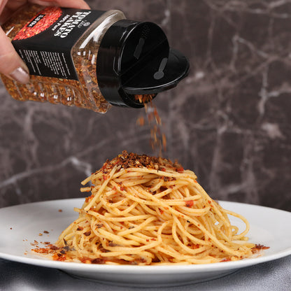 A hand sprinkles Ceres Gourmet Dried Tomato Powder, 100% pure, from a 5.3 oz shaker onto plain spaghetti on a white plate, against a marbled dark background. Perfect for sauces, soups, paste and more.