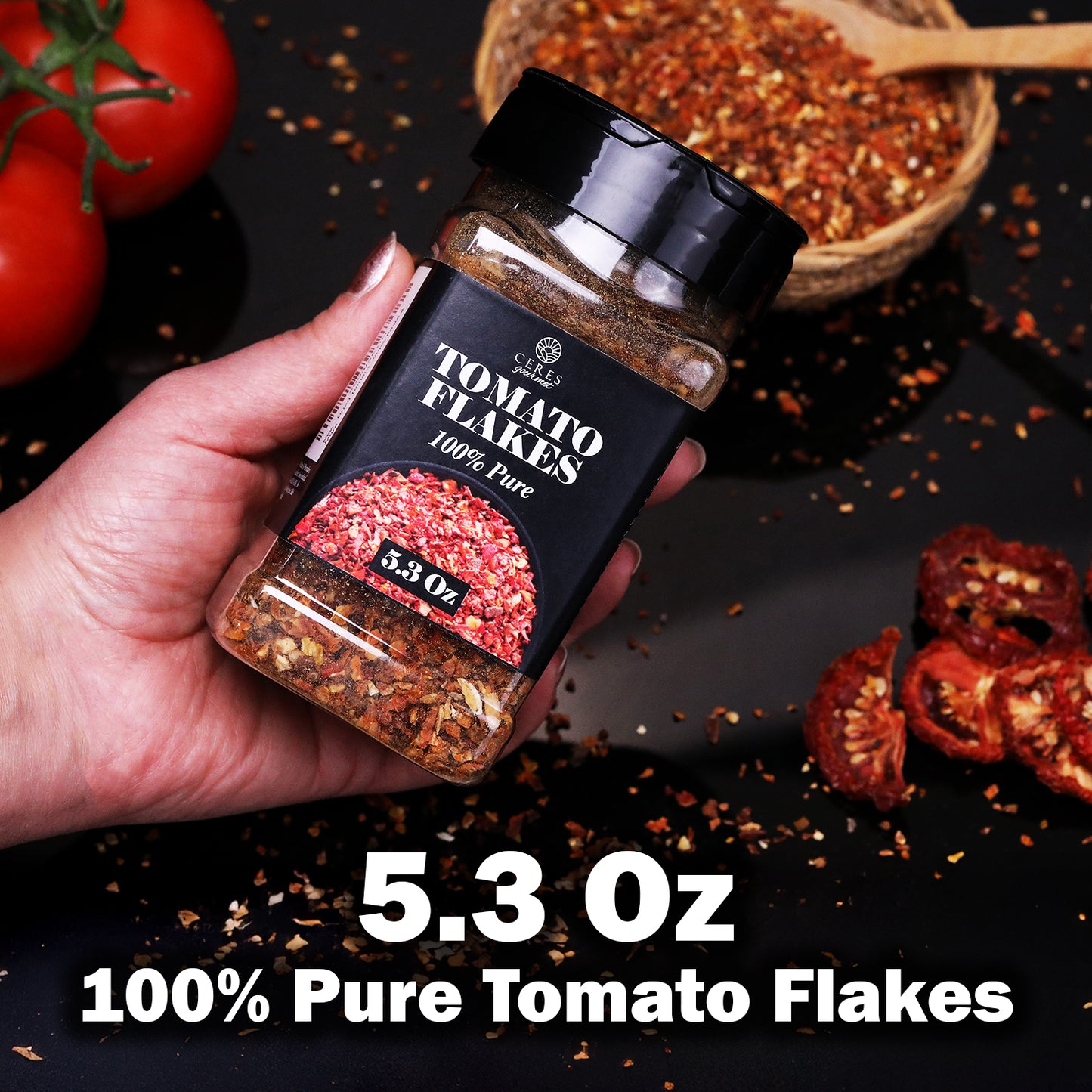 A hand holds a Ceres Gourmet Dried Tomato Powder jar (5.3 oz, 100% pure) above a dark surface with dried flakes, whole tomatoes, and a wooden bowl—ideal for sauces, soups, paste and more.