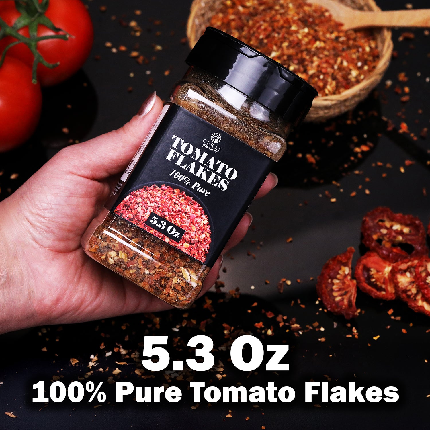 A hand holds a Ceres Gourmet Dried Tomato Powder jar (5.3 oz, 100% pure) above a dark surface with dried flakes, whole tomatoes, and a wooden bowl—ideal for sauces, soups, paste and more.