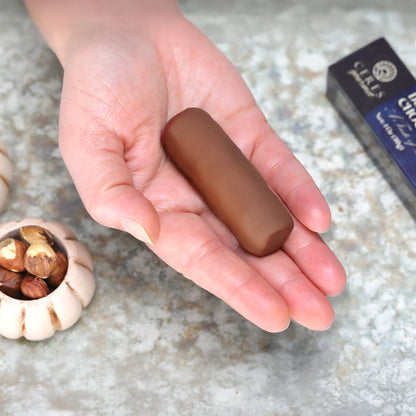 A hand holds a Ceres Gourmet Dubai Chocolate Hazelnut Mini Bar from the 3 Pack (1oz each), with hazelnuts and a chocolate box on a marbled surface, capturing the luxurious taste of Dubai-inspired chocolate treats.