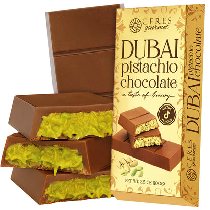 A Ceres Gourmet Dubai Chocolate Kataifi and Pistachio Gold Package stands upright beside chocolate pieces filled with green pistachio cream. The packaging displays images of the chocolates and pistachios, highlighting its Middle Eastern flavor.