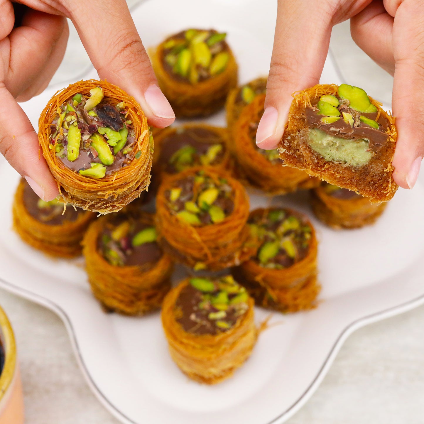 Close-up of hands holding Ceres Gourmet Dubai Chocolate Pistachio Baklava with Kataifi—a Middle Eastern dessert featuring crispy pastry, chocolate, and pistachios; more baklava is seen on a plate in the background.
