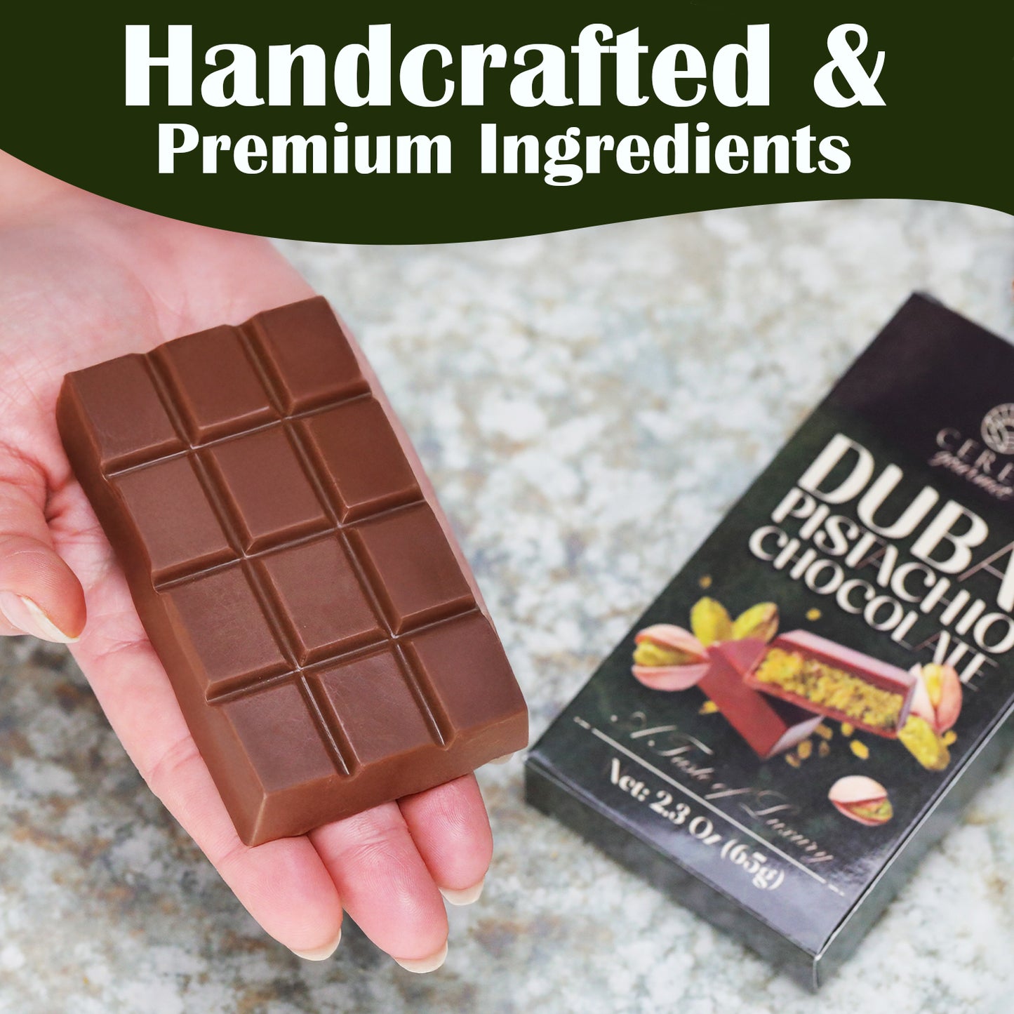 A hand holds a rectangular Ceres Gourmet Dubai Chocolate Bar (2.3 oz, 65g) beside its packaging on a marble surface. The box displays “Dubai Chocolate Bar” and above reads, “Handcrafted & Premium Ingredients.” Available in 3-12-24 Packs.