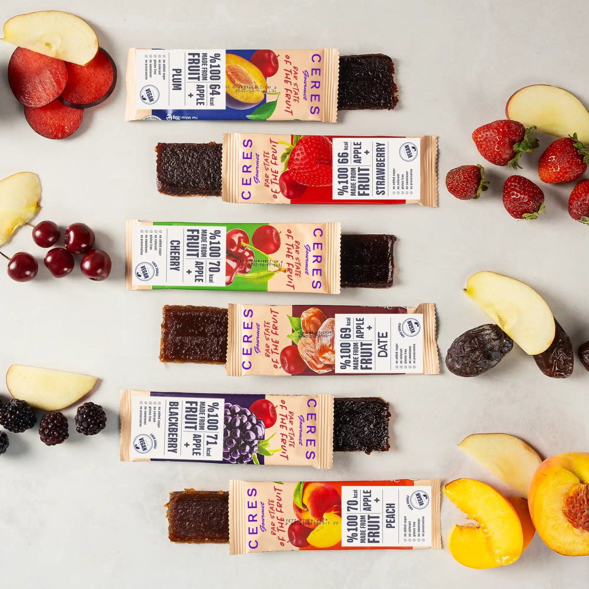 Five Ceres Gourmet Fruit Strips from the Variety Pack, each partly unwrapped, are lined up on a light surface. Fresh fruits—strawberries, apples, plums, grapes, and peaches—surround these vegan, gluten-free bars with no added sugar.
