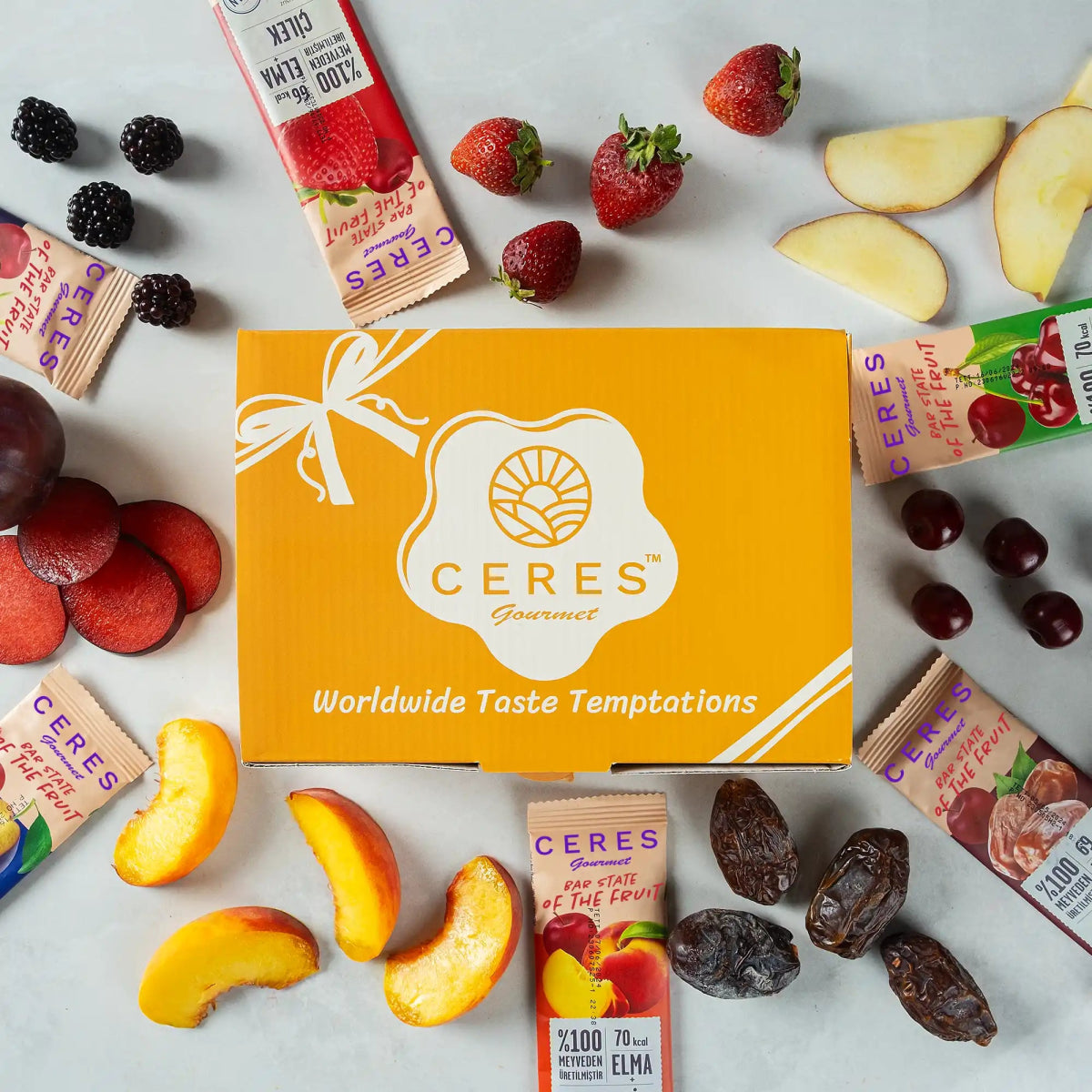 A yellow Ceres Gourmet box is surrounded by strawberries, blackberries, apple slices, grapes, peach slices, dates, plums, and Fruit Strips Variety Pack by Ceres Gourmet—vegan and gluten-free bars with no added sugar.