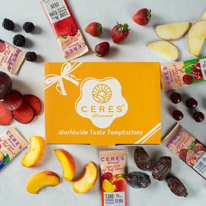 A yellow Ceres Gourmet box is surrounded by strawberries, blackberries, apple slices, grapes, peach slices, dates, plums, and Fruit Strips Variety Pack by Ceres Gourmet—vegan and gluten-free bars with no added sugar.