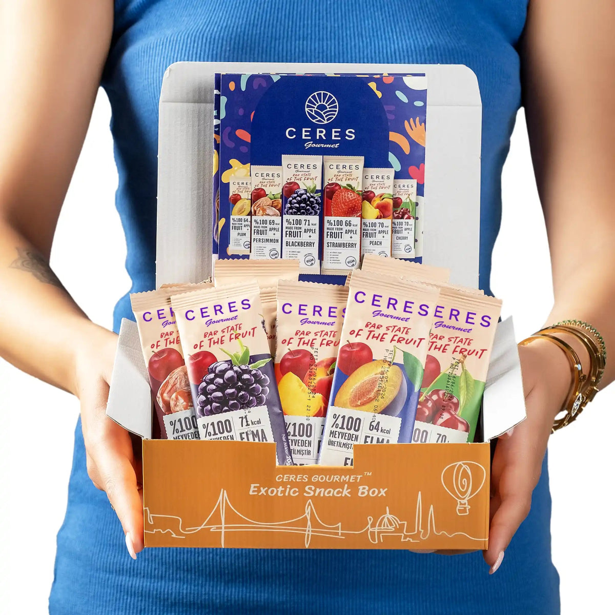 A person in a blue sleeveless top holds an open Fruit Strips Variety Pack by Ceres Gourmet, filled with vegan, gluten-free bars featuring colorful fruit strip packets and assorted fruits on the packaging, promoted as having no added sugar.