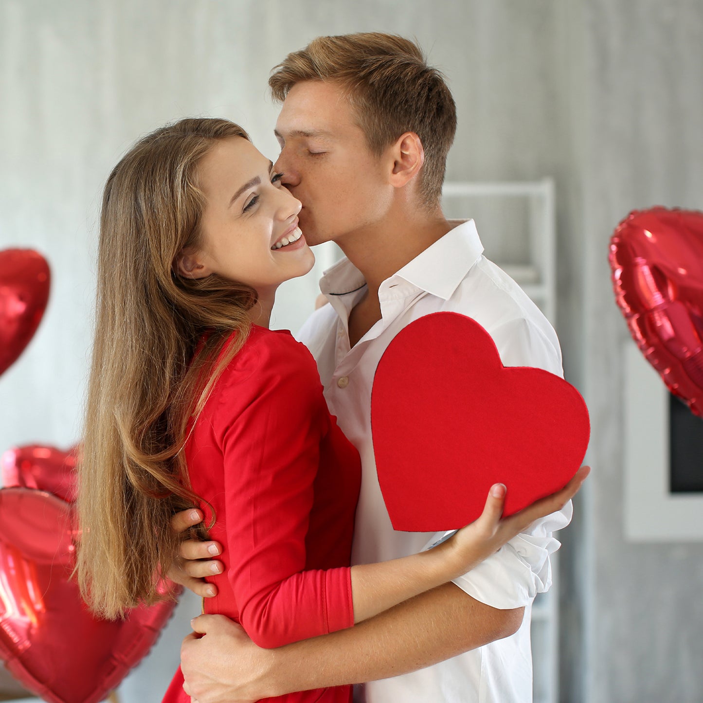 A smiling woman in a red dress holds a heart as her partner embraces her amid heart-shaped balloons—an ideal moment to gift the Ceres Gourmet Love Box International Snacks for Your Special Someone 💖 this Valentine’s Day.