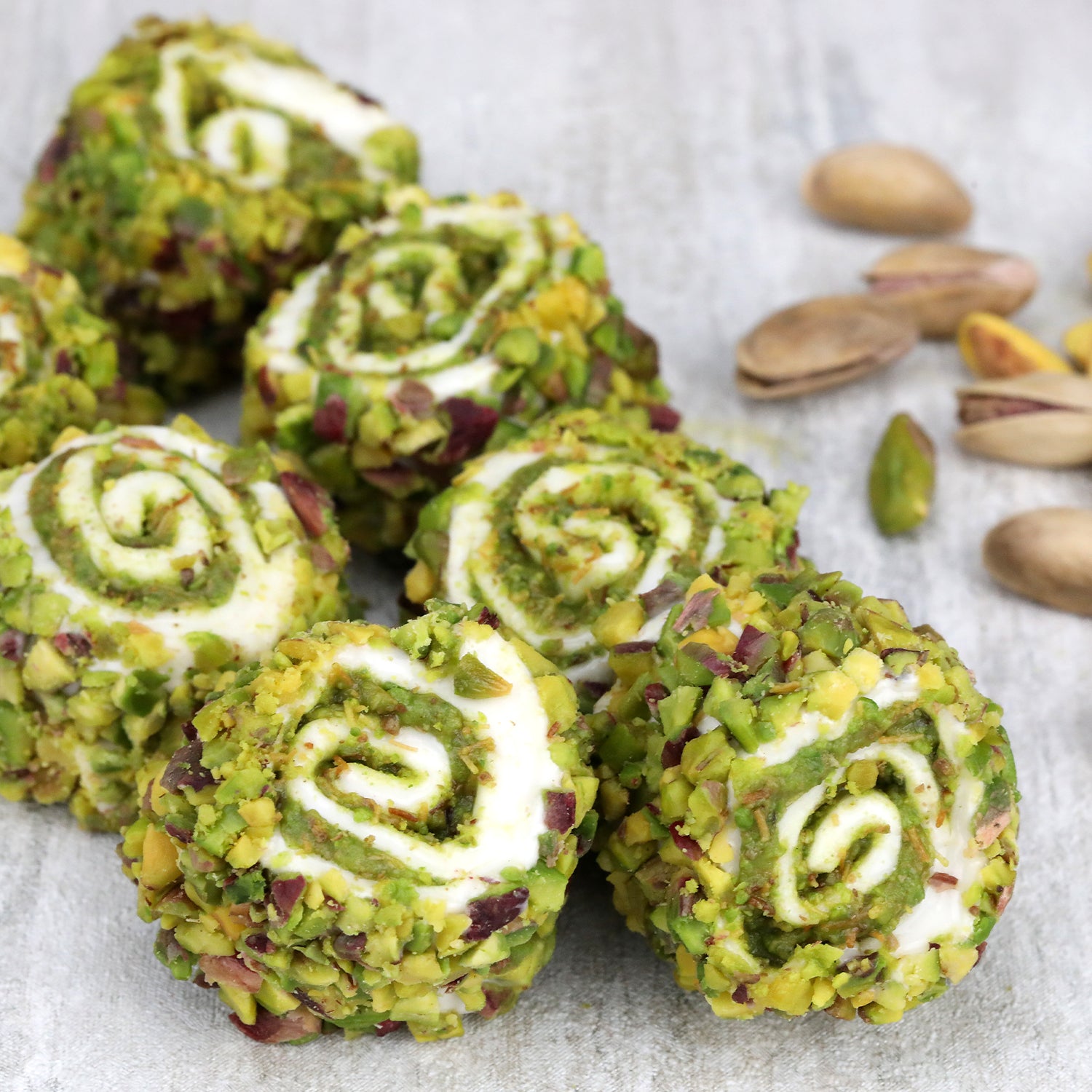 Ceres Gourmet’s Turkish Delight with Dubai Chocolate Filling (17.6 oz) features round, swirl-shaped sweets with a pistachio coating, arranged alongside whole and shelled pistachios on a light surface.