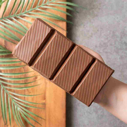A hand holds the Ceres Gourmet Viral Halloween Dubai Chocolate Bar, 75% Pistachio (7 oz)—a large, handcrafted treat with four segments and pistachio cream, set on wood with a palm leaf and gray backdrop.