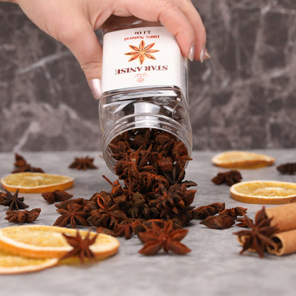 A hand pours Ceres Gourmet Whole Star Anise Pods - 2.1 oz from a resealable jar onto a gray surface with dried orange slices and cinnamon—ideal for baking, cooking, anise tea, and festive recipes.
